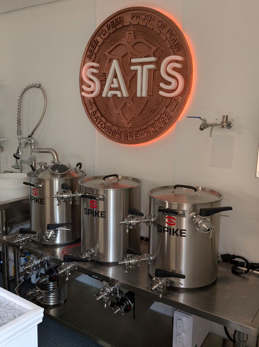 The satsbrew brewhouse: a lit copper SATS sign above three stainless steel Spike brew kettles